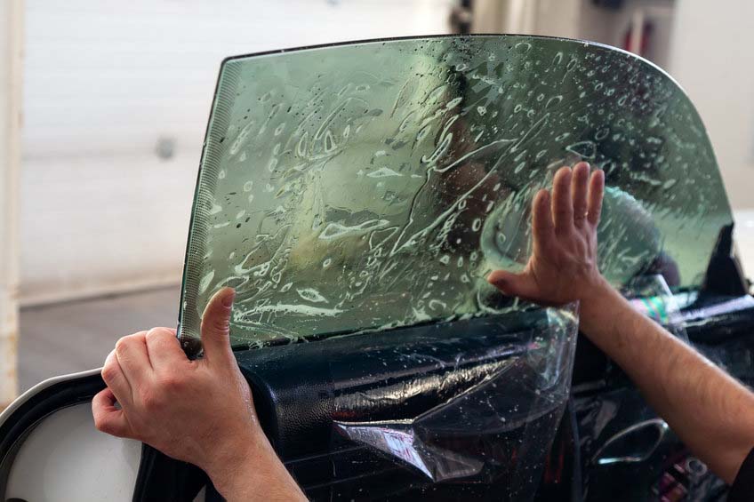 The wizard for installing additional equipment sticks a tint film on the side front glass of the car and flattens it by hand to fit the glass with a greenish tint in the auto service.